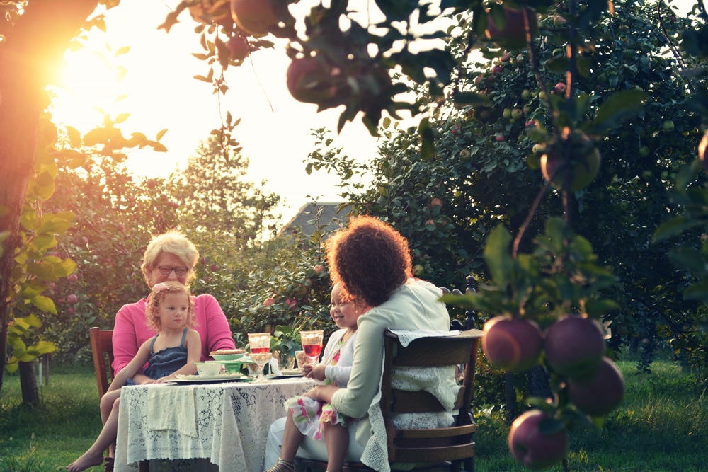 Zwei Erwachsene sitzen mit jeweils einem Kind auf dem Schoß an einem Tisch im Garten und unterhalten sich.